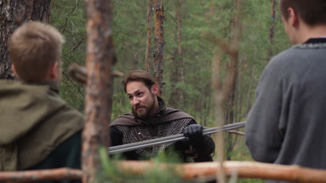 bearded man in armor with cape teaches children sword fight