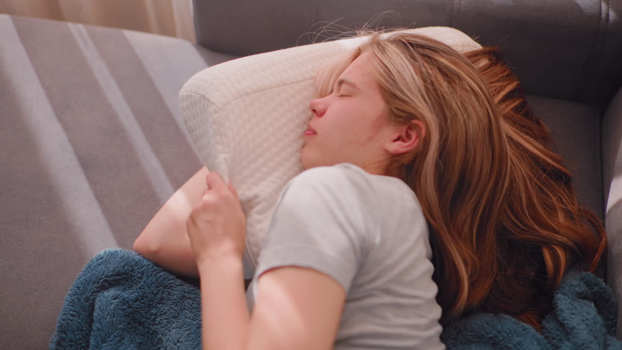 Top down view of exhausted woman lying on couch covering face with soft white pillow refusing to get up, eyes closed under morning light, wrapped in blue blanket seeking quiet from fatigue