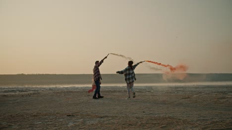 A-happy-middle-aged-man-in-a-checkered-shirt-together-with-his-little-daughter-and-his-wife,-a-brunette-woman-in-a-checkered-green-shirt,-are-holding-fireworks-in-their-hands-that-produce-green-and-red-smoke-during-their-relaxation-and-fun-outside-the-city-on-the-deserted-seashore-in-the-summer-even