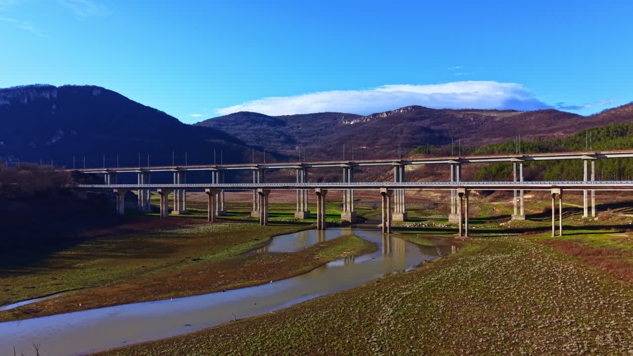 青空といくつかの雲が遠くに見える日中、丘に囲まれた川に架かる橋の建設