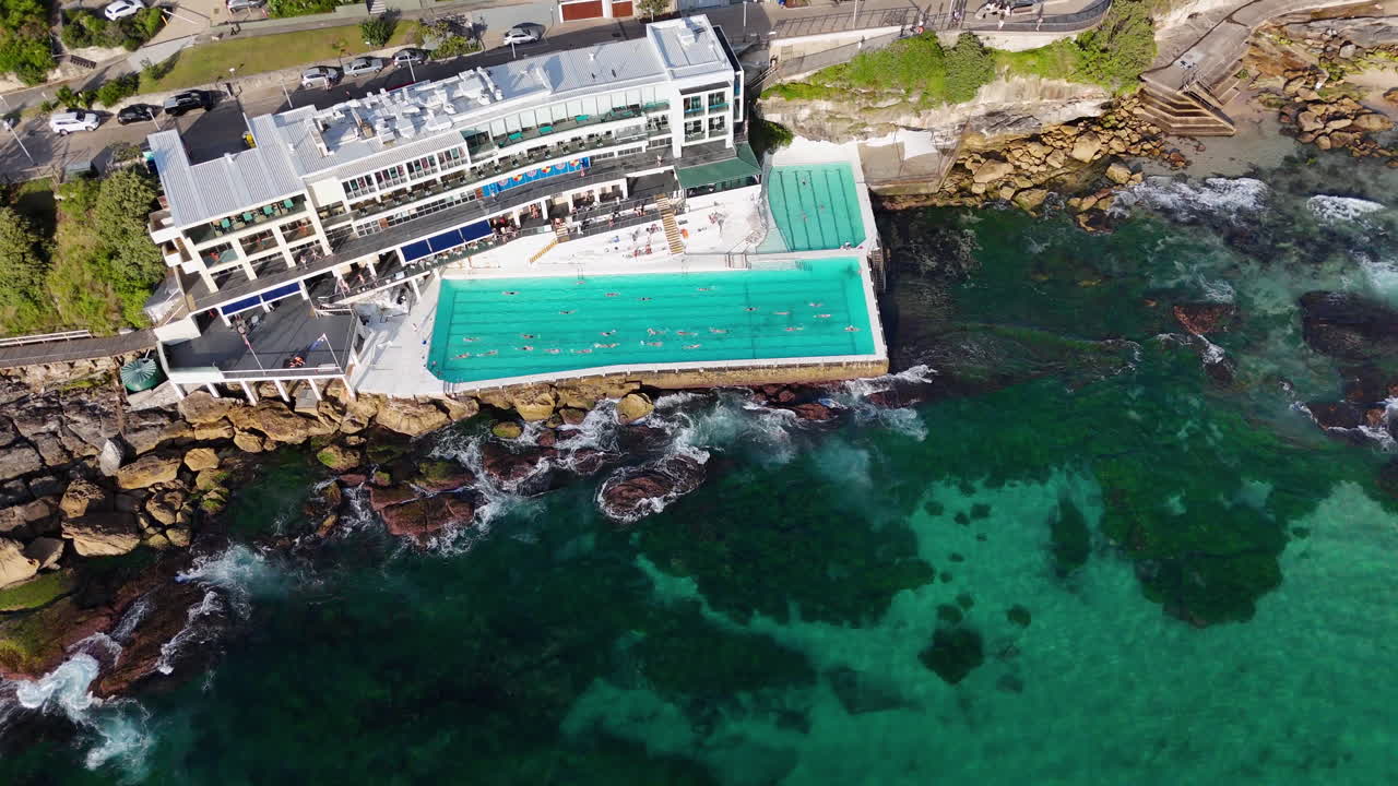 Drone footage of Bondi Beach, capturing the ocean pool nestled against the coastline, with people swimming and relaxing. Downward angle aerial footage.