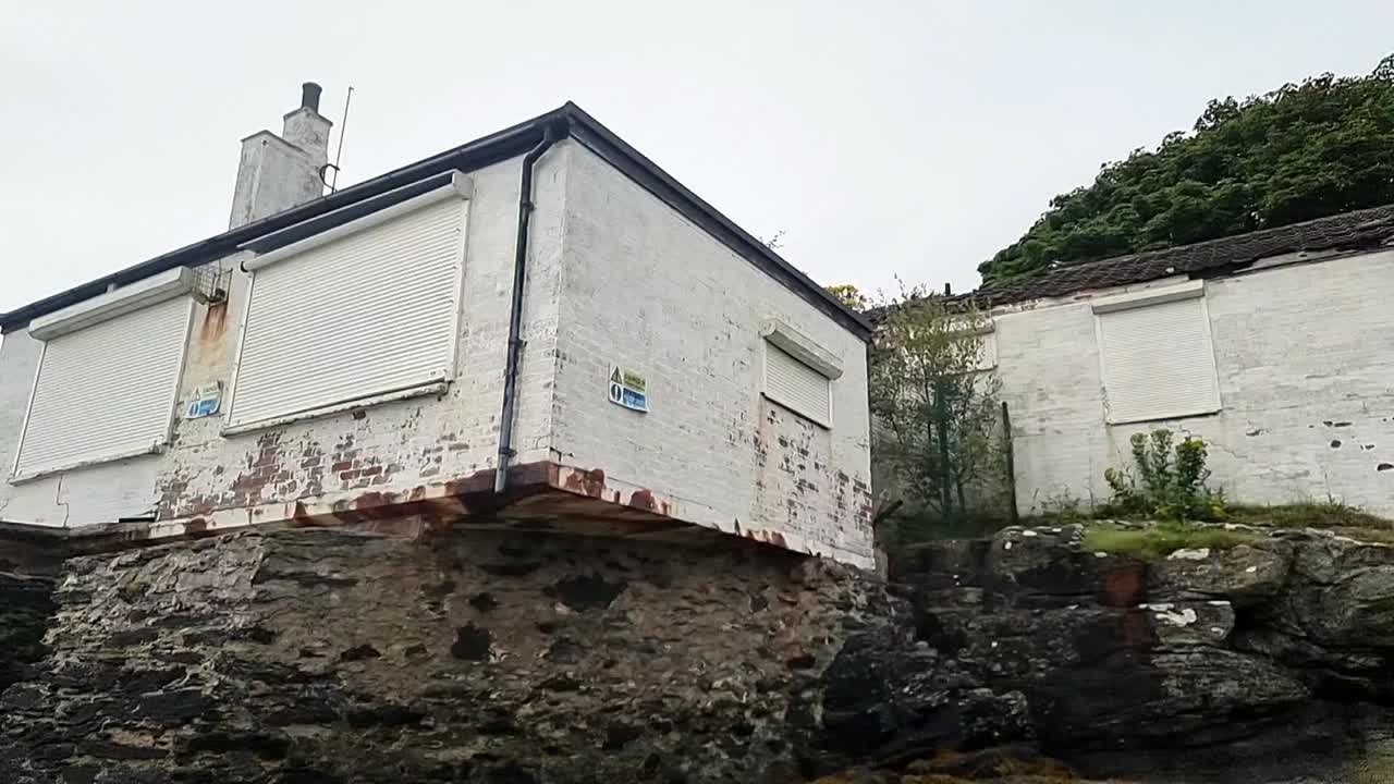 Low-angle shot of a decaying white house on the beach in Penrhos. The closed shutters and cracked base suggest abandonment and coastal damage
