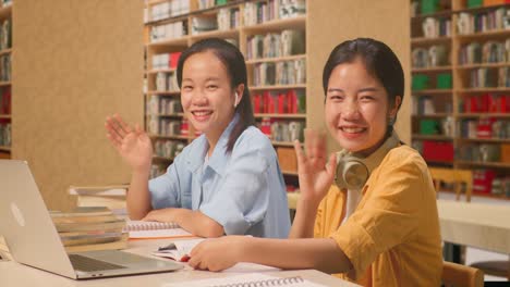 asian woman students with headphones smiling to camera and waving hands while reading books on the table with laptop in the library