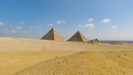 Low-pan-across-desert-sands-with-Great-Pyramids-of-Giza-on-distance-under-clouds