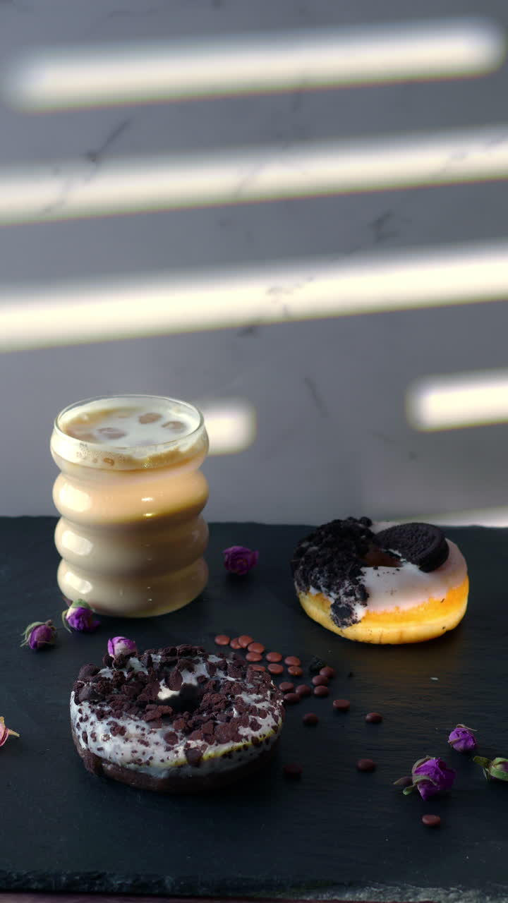 A hand with pink nails reaches for a donut topped with cookie crumbles, beside iced coffee on a slate surface. Dried rosebuds and chocolate drops complete the soft-lit cozy setting.