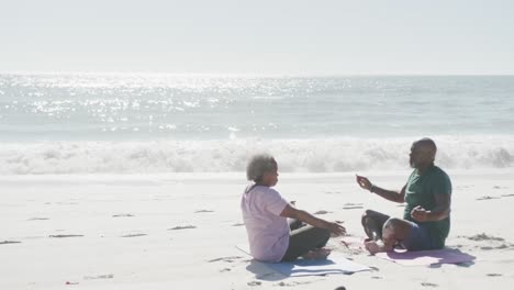 happy senior african american couple doing yoga and meditating at beach, copy space, in slow motion