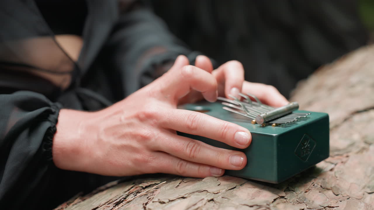 Close up of hands in dark outfit gently plucking metal tines on small green thumb piano resting on tree bark, surrounded by natural outdoor forest light, evoking calm emotion