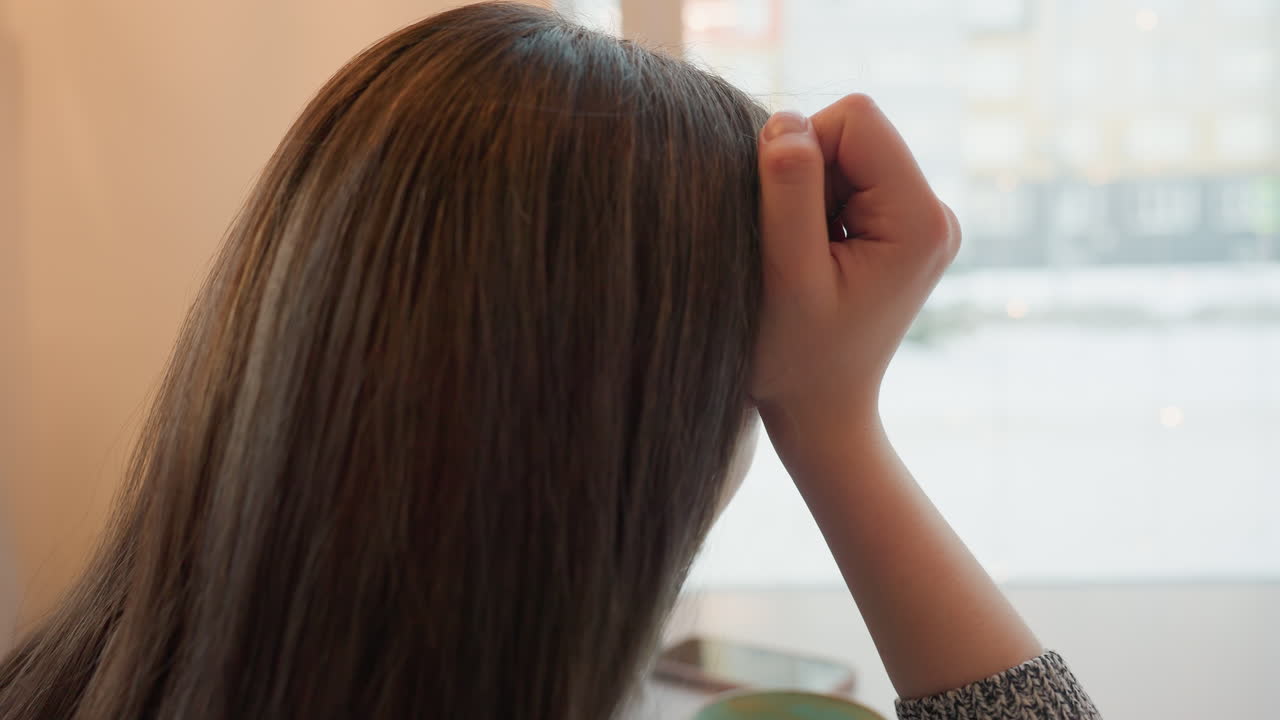 Young woman with long straight hair sits quietly indoors resting head on hand while thoughtfully staring outside cafe window during snowy day with coffee cup and mobile phone on table beside her