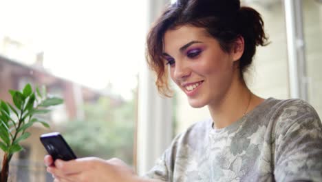 Mujer-Joven-Y-Atractiva-Enviando-Mensajes-De-Texto-En-Su-Teléfono-Móvil-En-La-Cafetería-Y-Sonriendo.-Mujer-Usando-La-Aplicación-En-El-Teléfono-Inteligente-En-La-Cafetería-Tomando-Café