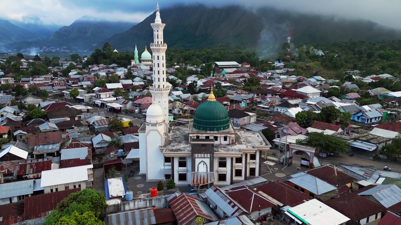 Desa pedesaan yang berpusat di sekitar Masjid Islam di bawah latar belakang pegunungan yang dramatis, dengan rumah-rumah berkelompok tersebar di lembah gunung yang subur, rekaman drone udara.