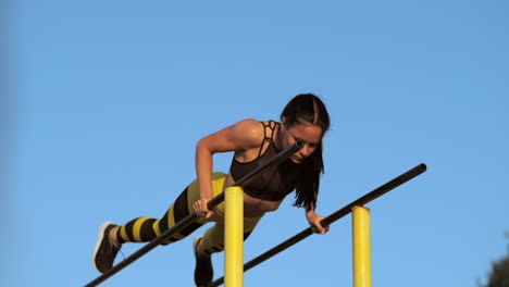 una hermosa atleta mujer delgada con una camiseta negra y pantalones amarillos al atardecer realiza flexiones en una barra horizontal paralela en cámara lenta.