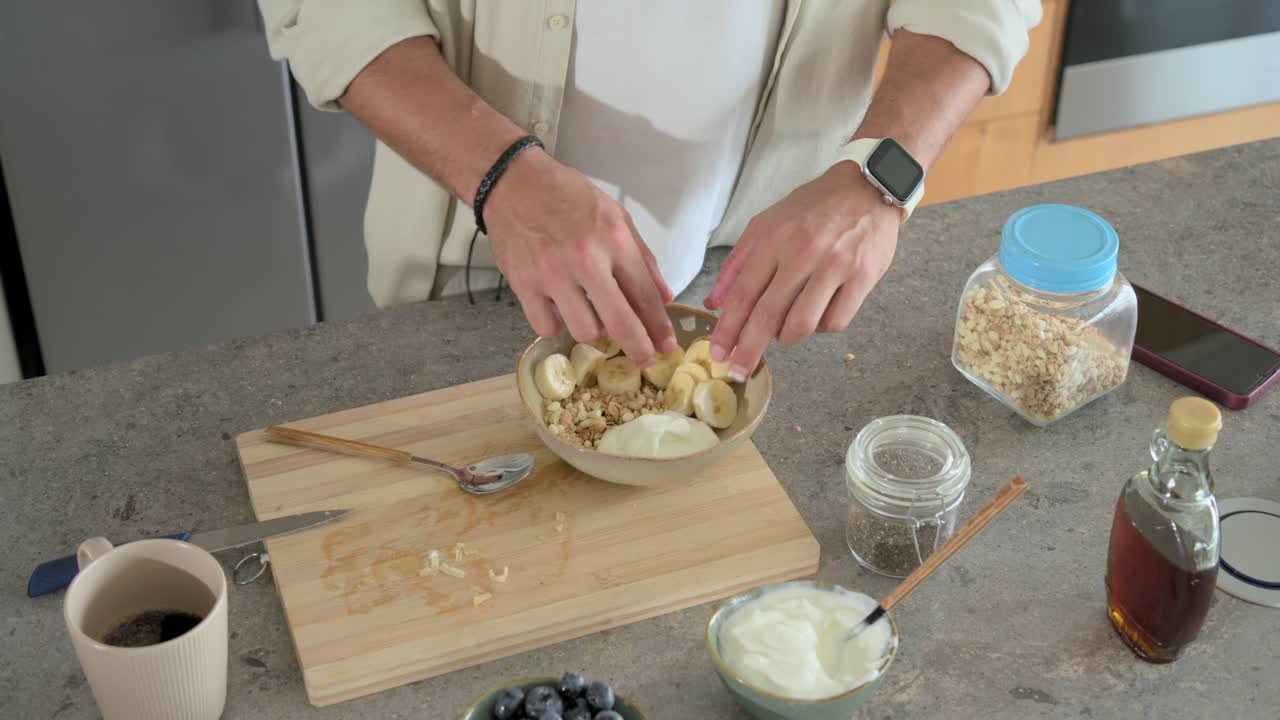Mand skærer banan på skærebræt, flytter skiverne ned i en granolaskål og tilføjer yoghurt til morgenmad.