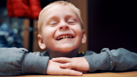 young little blond boy is looking at the camera. the child is shaking his head with different expressions on his face on the blurred background. happy boy sitting in the room. close-up