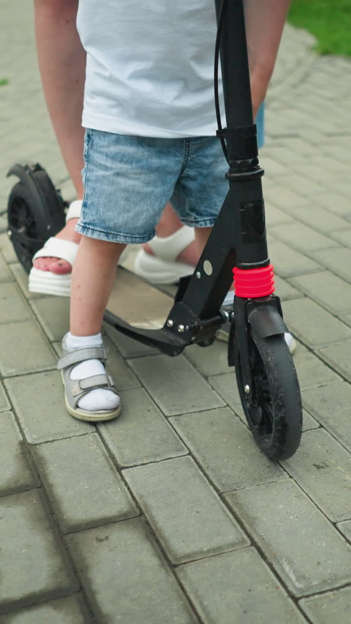 A close-up leg view of a child and woman standing on a scooter along an interlocked path outdoors