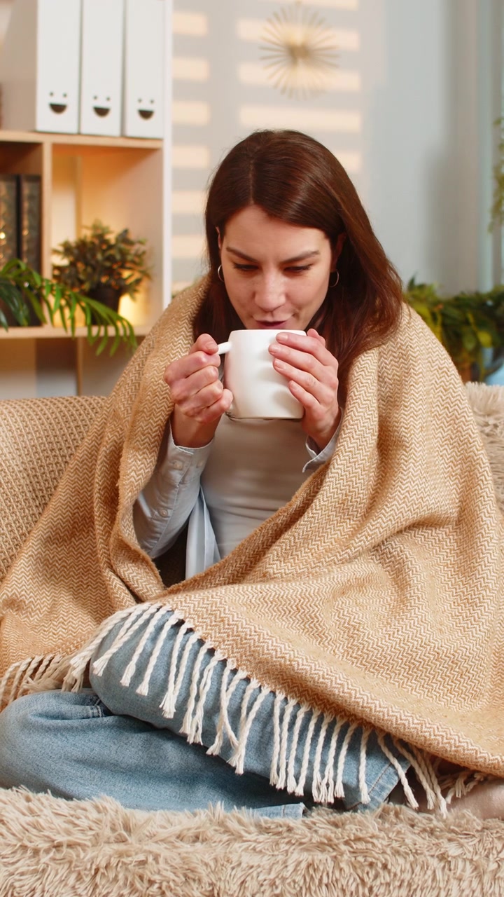 Giovane donna sul divano di casa che si riscalda con una bevanda calda sotto una coperta, godendosi un tranquillo relax casalingo al chiuso.