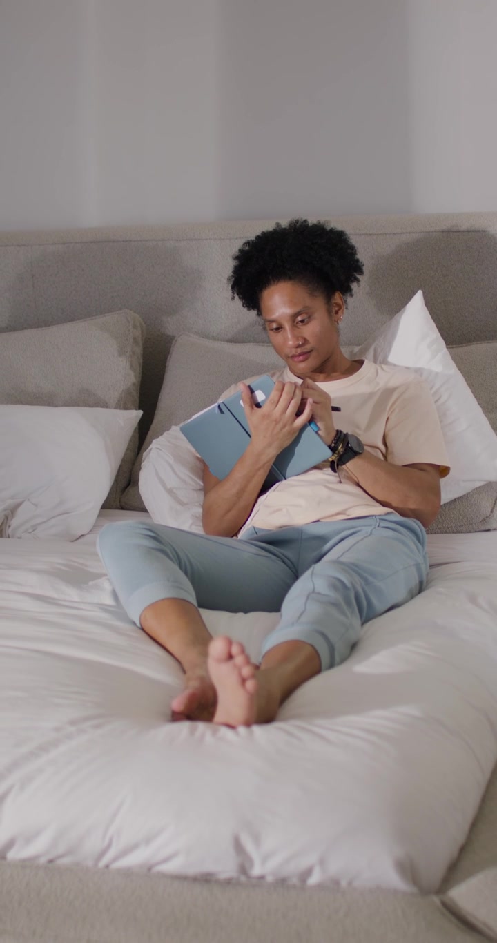 Video verticale: Uomo afroamericano che porta un tablet blu sui cuscini, che tocca lo schermo e legge testi a letto.