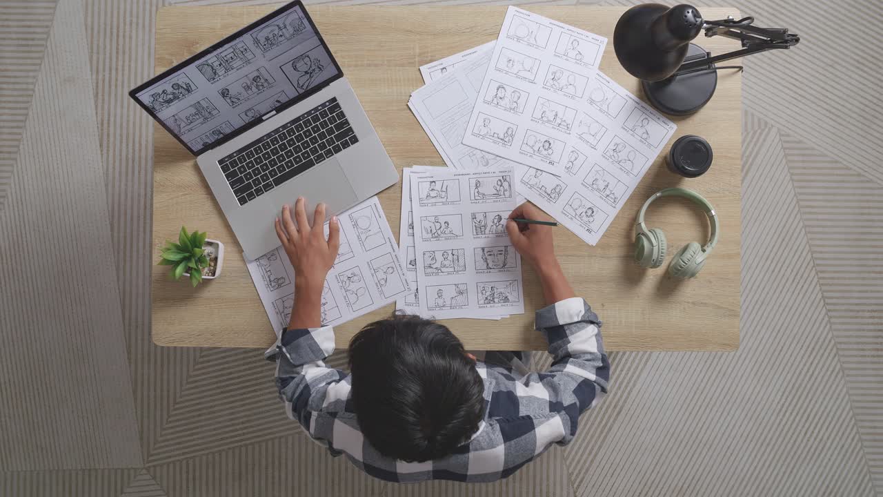 Top View Of Asian Male Artist Looking At Sketching Concept On A Laptop Then Drawing Storyboard On The Table With Headphones In The Studio