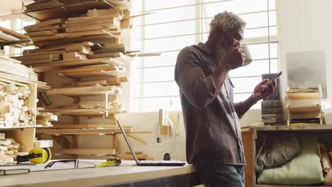 african american male carpenter using smartphone while drinking coffee in a carpentry shop