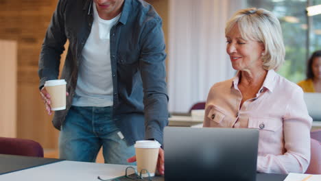 Reife-Geschäftsfrau-Betreut-Jüngeren-Männlichen-Kollegen,-Der-Am-Laptop-Am-Schreibtisch-Arbeitet