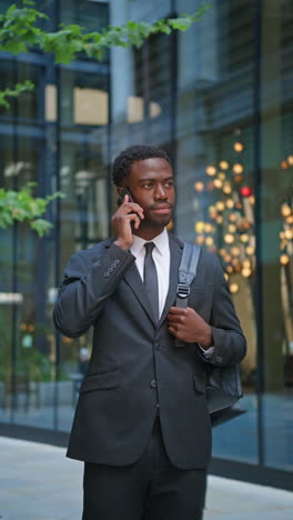 Vertical-Video-Shot-Of-Smiling-Young-Businessman-Wearing-Suit-Using-Mobile-Phone-Walking-Outside-Past-Offices-In-The-Financial-District-Of-The-City-Of-London-UK-Shot-In-Real-Time