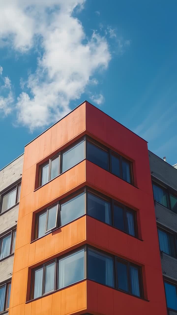 Vídeo vertical: la cámara se inclina hacia arriba desde la esquina de un edificio naranja, pasando por una fachada gris blanquecina hacia un cielo azul