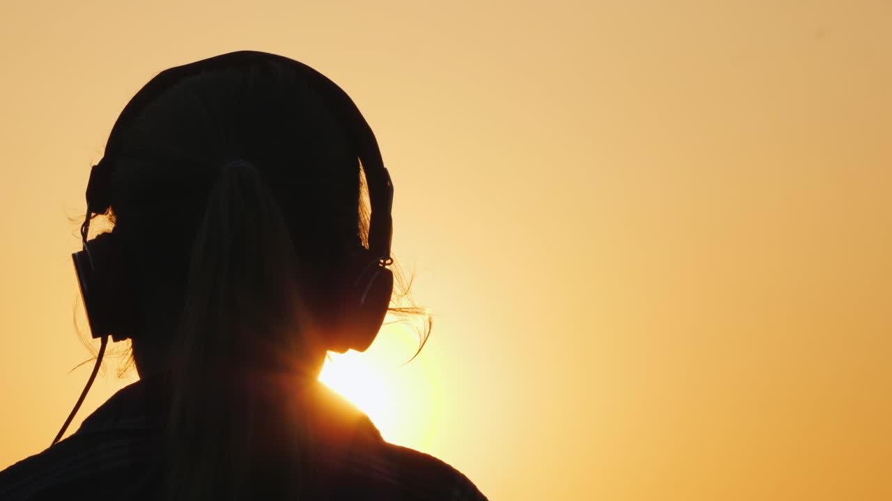 silueta de una chica con auriculares escuchando música en el telón de fondo de un gran sol poniente y cielo naranja
