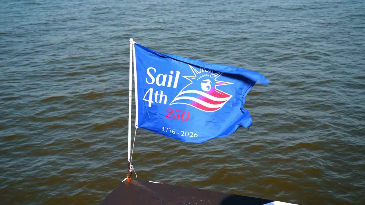 Bandiera commemorativa del 4° Sail che sventola sopra l'acqua del porto. Lo striscione blu ondeggia nella brezza sopra la superficie scura e increspata di un fiume in un pomeriggio soleggiato