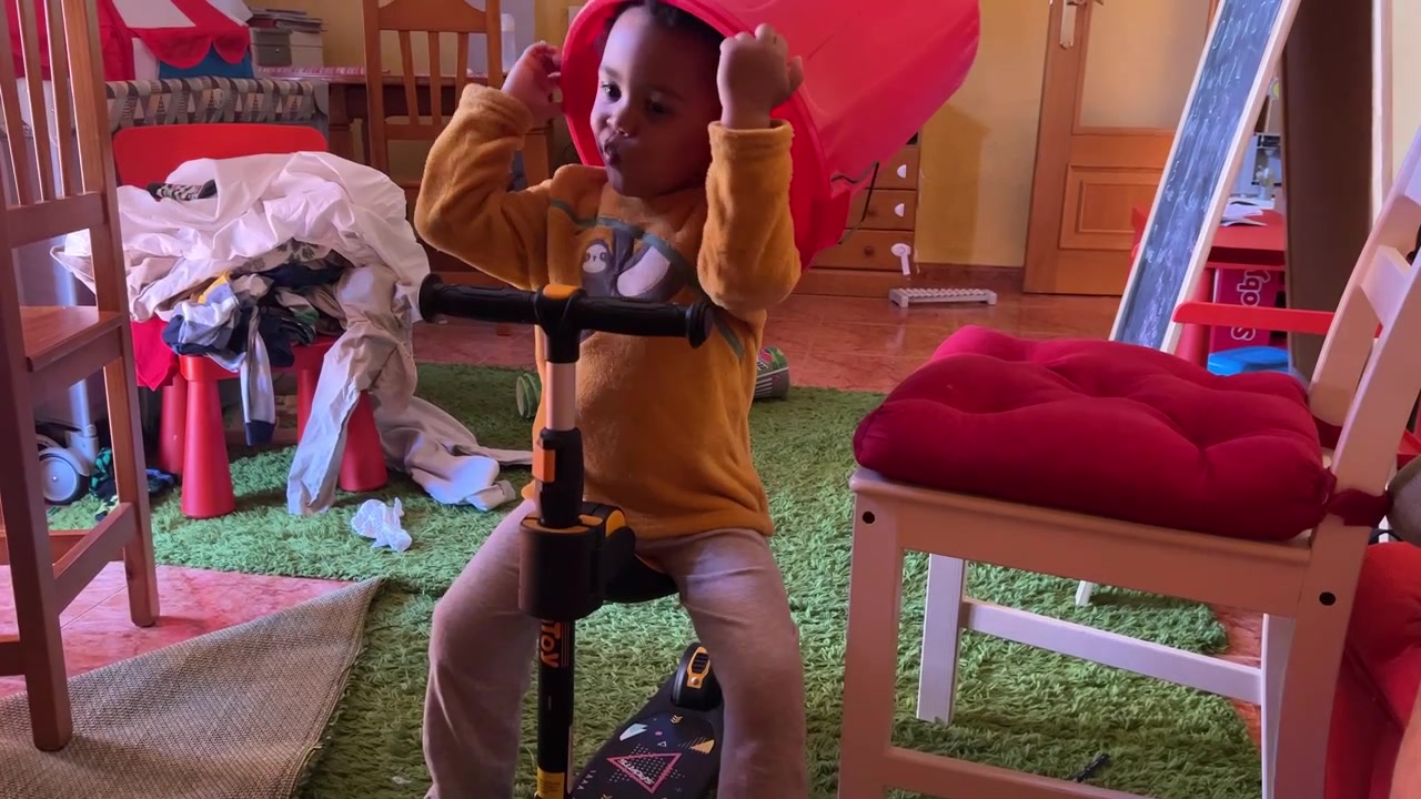 Three-year-old funny black child playing at home with laundry basket while riding his scooter