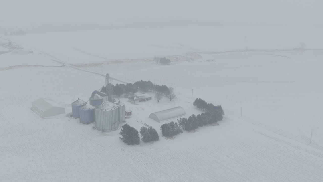 Aerial pan shot of farmstead with several large silos in a snowy landscape of blizzard