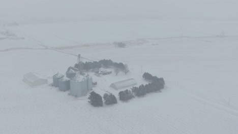 aerial pan shot of farmstead with several large silos in a snowy landscape of blizzard