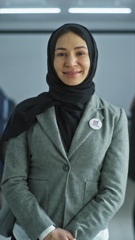 Portrait-of-mature-woman,-United-States-of-America-elections-voter.-Businesswoman-stands-in-a-modern-polling-station,-poses-and-looks-at-camera.-Background-with-voting-booths.-Concept-of-civic-duty.