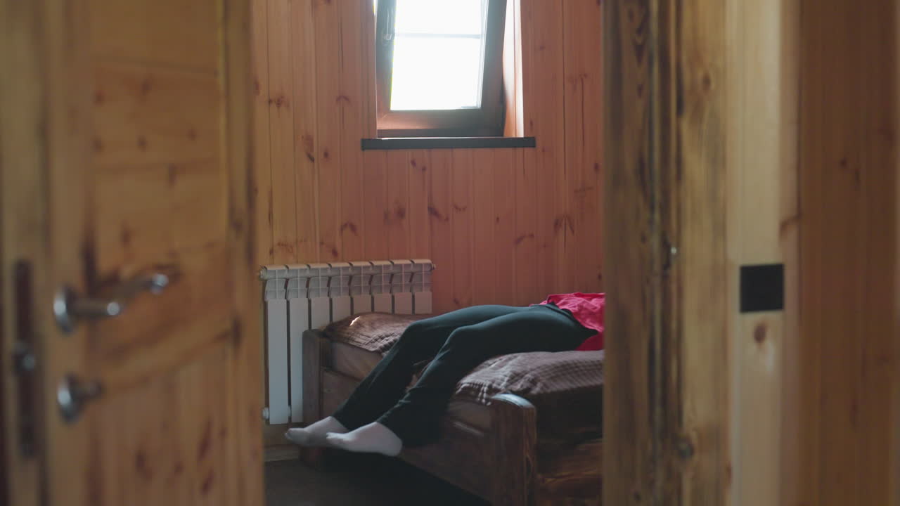 young lady wearing red shirt enters cozy wooden room and walks toward bed to sit down calmly, soft light streaming through window creates peaceful indoor atmosphere with sense of relaxation