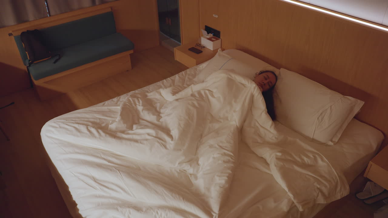 호텔 침대에서 뒤척이는 여성 투숙객, 흐트러진 하얀 이불, 희미한 침대 옆 램프 불빛, 이불 속에서 뒤척임, 불면증과 불안, 위에서 본 시점, 부티크 호텔 객실, 밤 분위기, 혼자