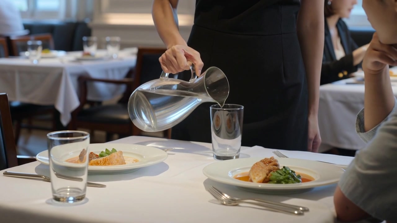 Walking server in fitted black uniform holding pitcher, pouring water to refill glass at restaurant