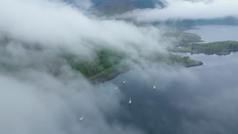 Aerial-tilt-down-flying-through-clouds-and-revealing-boats-on-coast-of-Glen-Coe,-Scotland