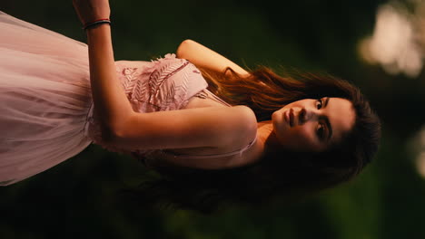 young caucasian beautiful girl in pink dress stares at camera, plays with hair at botanical garden, outdoors, vertical video