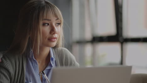 A-young-Asian-girl-looks-out-the-window-and-thinks-sitting-with-a-laptop-at-her-desk.