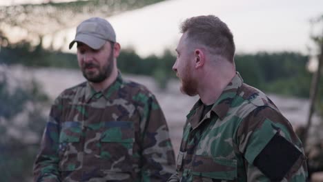 soldiers in camouflage clothing, discussing in the evening at a common bonfire frying sausages for supper, men joking, laughing, resting after work, service, field exercises