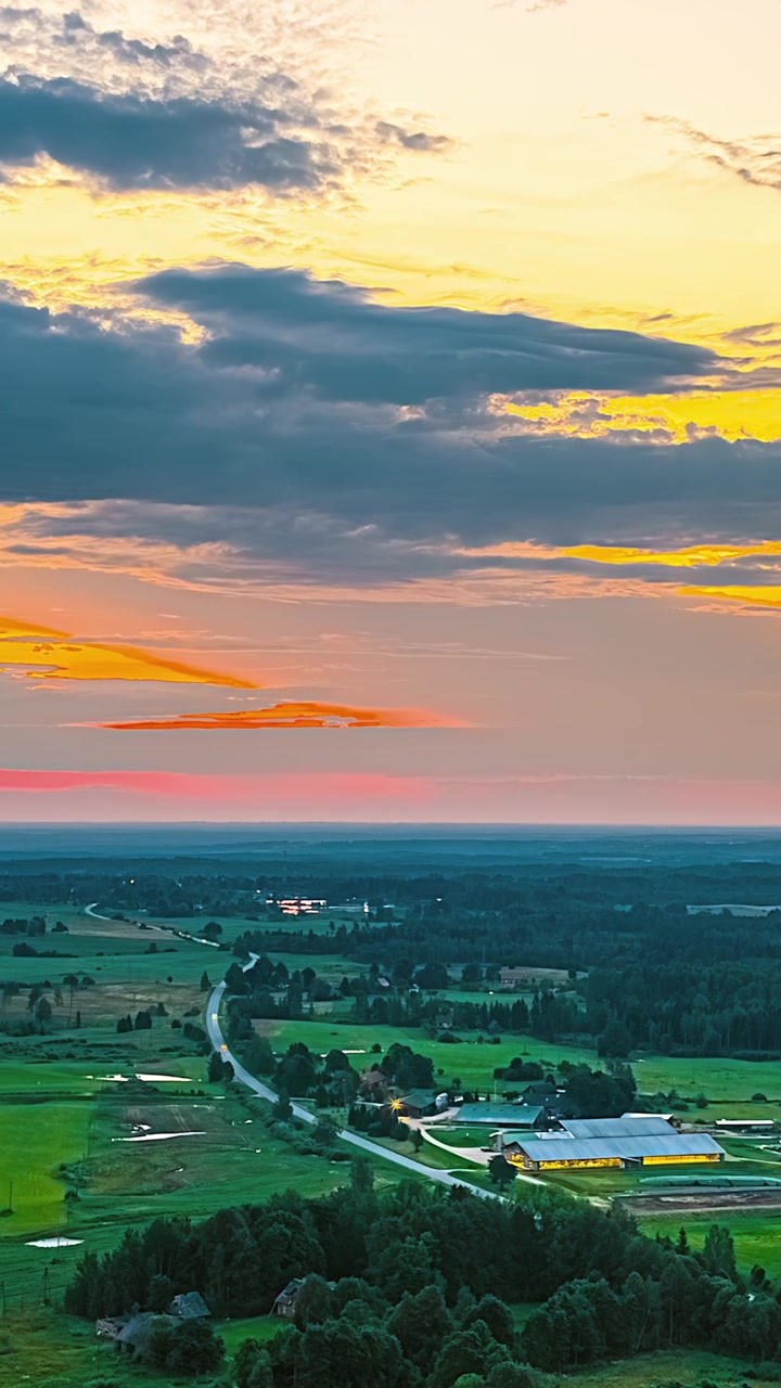 Una strada tortuosa si snoda attraverso la lussureggiante campagna lettone verso una fattoria illuminata sotto un cielo drammatico al tramonto - time-lapse verticale aereo