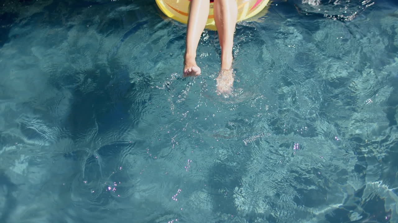Teenage girl''s legs dangle from a yellow float into clear blue pool water