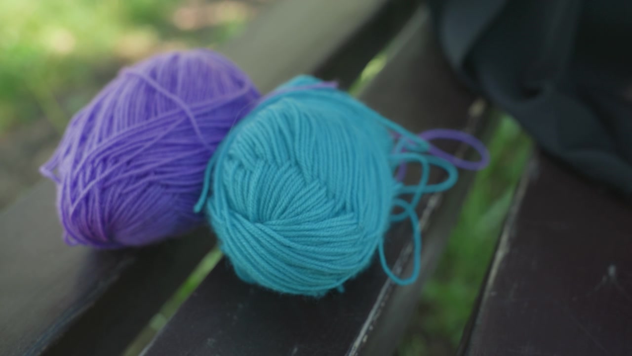 Close up of three balls of yarn in purple turquoise and white on wooden bench. hand briefly moves ball of white yarn before removing it from view. Soft natural light illuminates scene