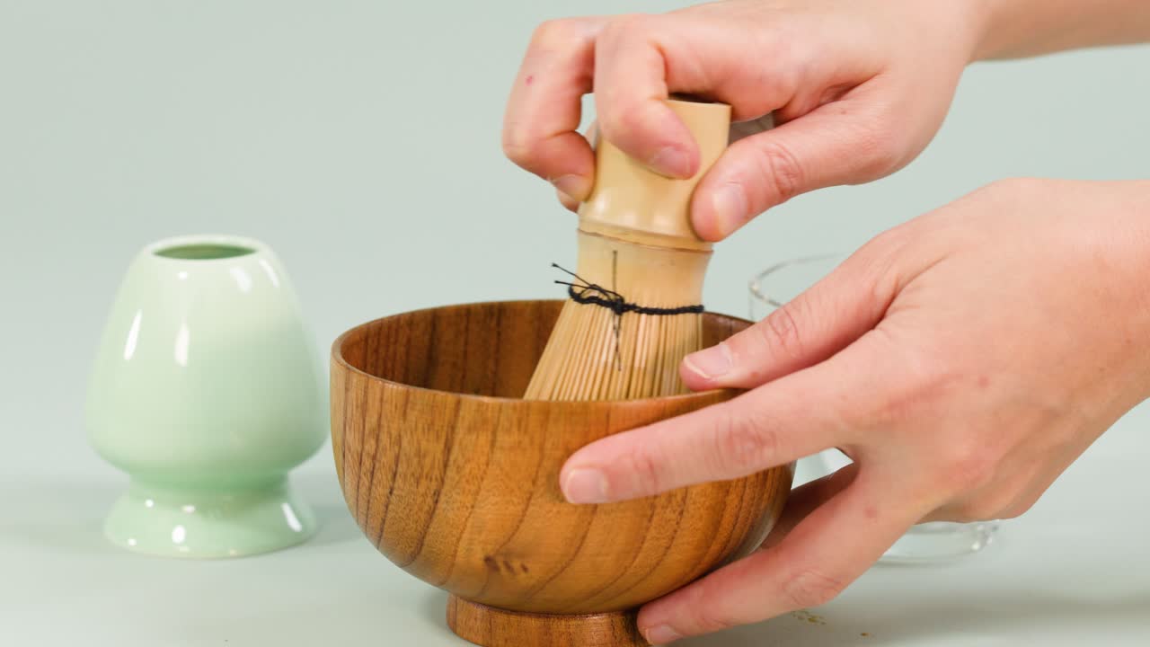 Hands whisk matcha in wooden bowl using bamboo whisk, minimal clean background, soft natural lighting