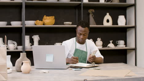 male small business owner counting money and writing on clipboard in the pottery shop