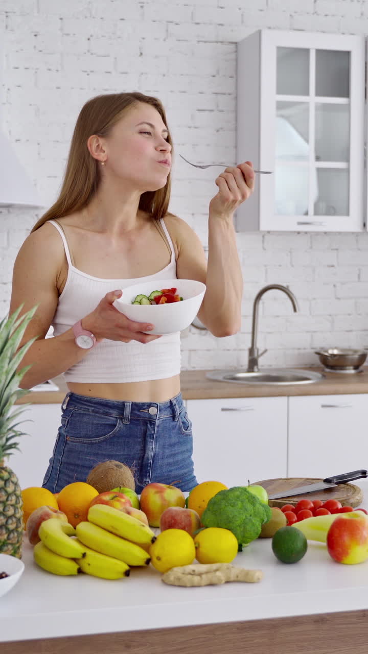 Young woman in the kitchen. Beautiful girl standing near the table with organic food. Lovely female enjoys eating fresh salad on the modern kitchen background. Dieting concept. Vertical video
