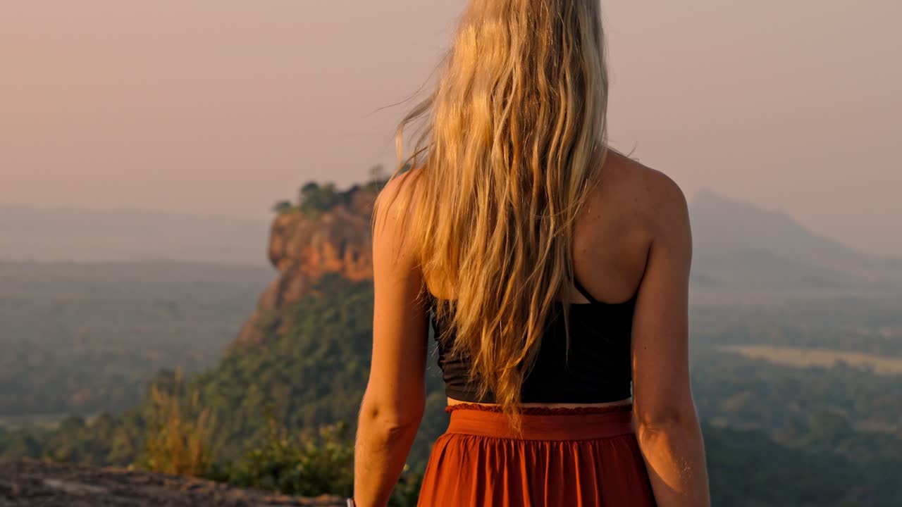 A woman walks peacefully with the majestic Sigiriya Rock landmark visible in the background during a vibrant sunrise in Sri Lanka.