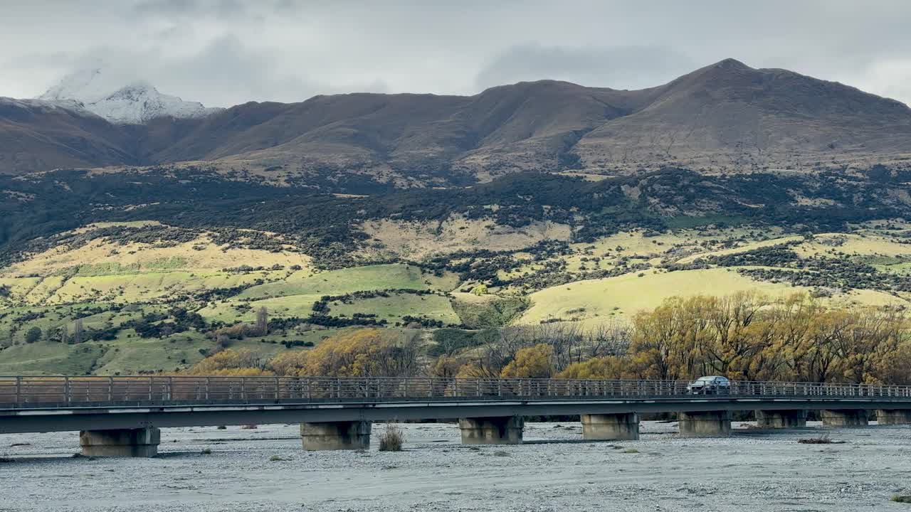 A truck drives across a bridge spanning a river, with mountains and cloudy sky behind