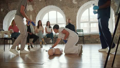 close-up shooting: a bearded man brunette in a gray t-shirt starts a practical lesson showing first aid together with a professional doctor a woman in a white uniform with a mannequin in front of the public against the background of brick walls and large windows