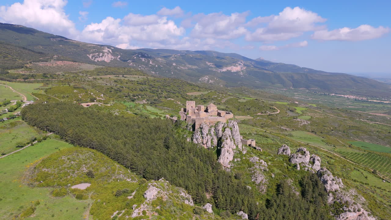 Lateral drone flight showcasing a medieval castle perched on a rocky hill, surrounded by dense forest and expansive rural landscape under a blue sky with scattered clouds. Scenic and historic view