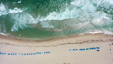 Aerial-drone-shot-looking-straight-down-flying-right-over-umbrellas-and-tourists-tanning-on-a-beach-while-blue-water-waves-crash-towards-them-on-Singer-Island-in-West-Palm-Beach-Florida