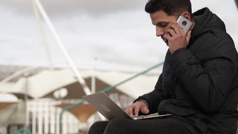 Concentrated-young-man-talking-on-phone-and-using--laptop